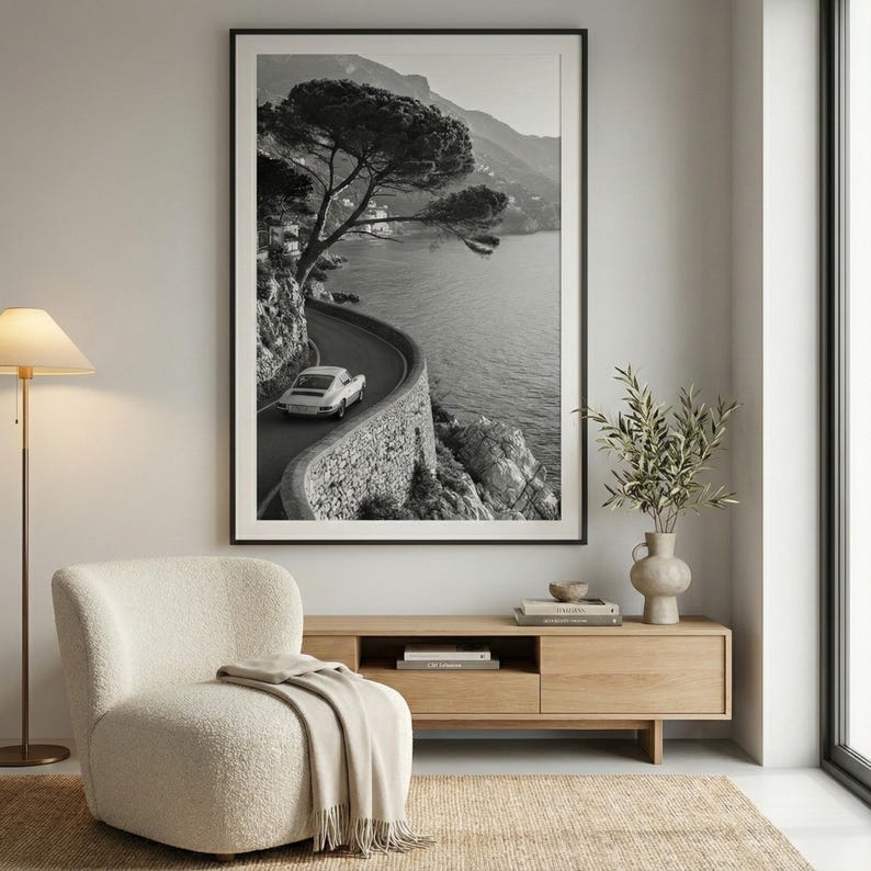 Op de afbeelding: Zwart-wit ingelijste print van een kustweg met een oldtimer. De kronkelende weg volgt de kustlijn, met een stenen muur en de zee. Een boom hangt over de weg. De print wordt boven een houten kast weergegeven.