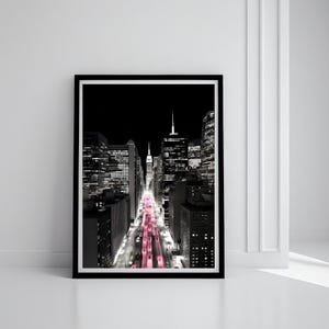 May include: Black and white framed print of a city street at night. The street lights are a vibrant pink, contrasting with the monochrome buildings. The artwork is in a black frame and is displayed against a white wall.