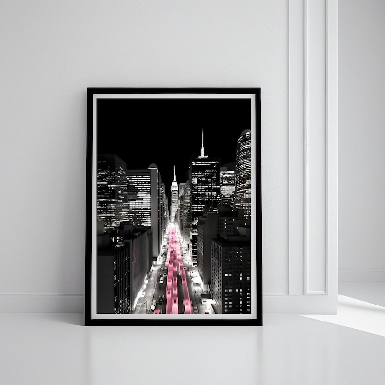 May include: Black and white framed print of a city street at night. The street lights are a vibrant pink, contrasting with the monochrome buildings. The artwork is in a black frame and is displayed against a white wall.