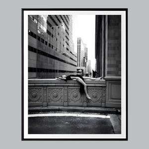 Pode incluir: Uma fotografia a preto e branco de uma pessoa com um vestido escuro deitada horizontalmente sobre um corrimão de pedra ornamentado. O fundo exibe um arranha-céus urbano com janelas de vidro e detalhes arquitetónicos, dentro de uma moldura.
