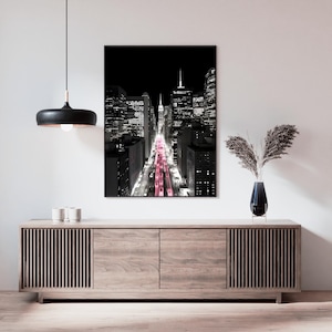 May include: Black and white cityscape art print with a black frame. The image features a view down a city street with pink lights. The print is displayed above a wooden cabinet with a black lamp and vase.