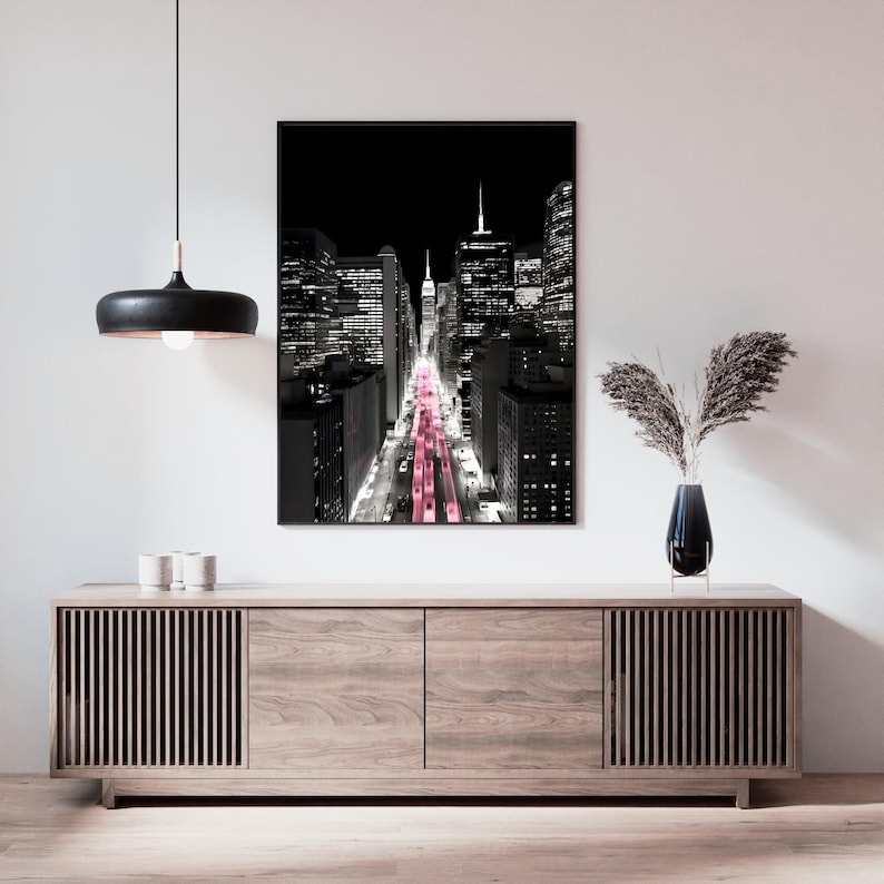 May include: Black and white cityscape art print with a black frame. The image features a view down a city street with pink lights. The print is displayed above a wooden cabinet with a black lamp and vase.
