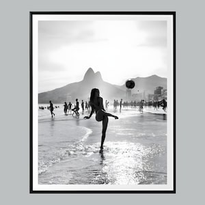 May include: Black and white photograph of a woman kicking a soccer ball on a beach. Silhouetted figures play in the background, with mountains and a city skyline visible. The image is framed in a black border.