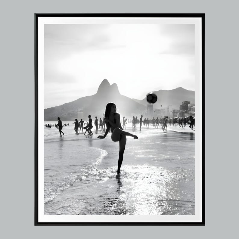 May include: Black and white photograph of a woman kicking a soccer ball on a beach. Silhouetted figures play in the background, with mountains and a city skyline visible. The image is framed in a black border.