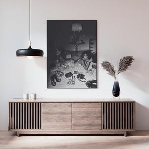 Op de afbeelding: Een zwart-wit ingelijste print van een vrouw omringd door schoenen en handtassen. De print hangt boven een licht houten kast met verticale lamellen. Een zwarte hanglamp hangt aan het plafond.