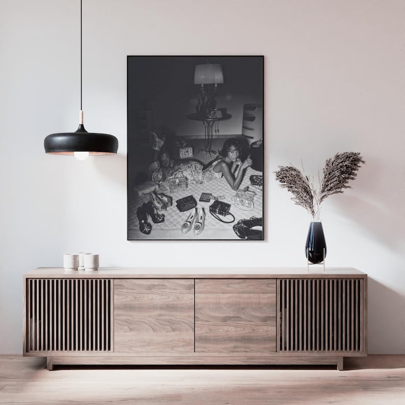 Op de afbeelding: Een zwart-wit ingelijste print van een vrouw omringd door schoenen en handtassen. De print hangt boven een licht houten kast met verticale lamellen. Een zwarte hanglamp hangt aan het plafond.