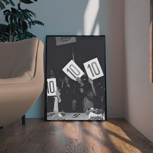 May include: Black and white framed print of a vintage event. People are holding up signs with the number "10" on them. A sign for "Sterling Vodka" is visible at the top. The image is displayed on a wooden floor.