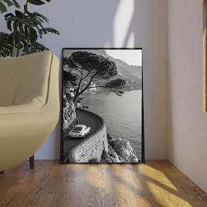 Op de afbeelding: Zwart-wit ingelijste print van een kustweg met een witte auto. De weg slingert langs de kustlijn, met aan de ene kant een stenen muur en aan de andere kant de zee. Een grote boom hangt over de weg.