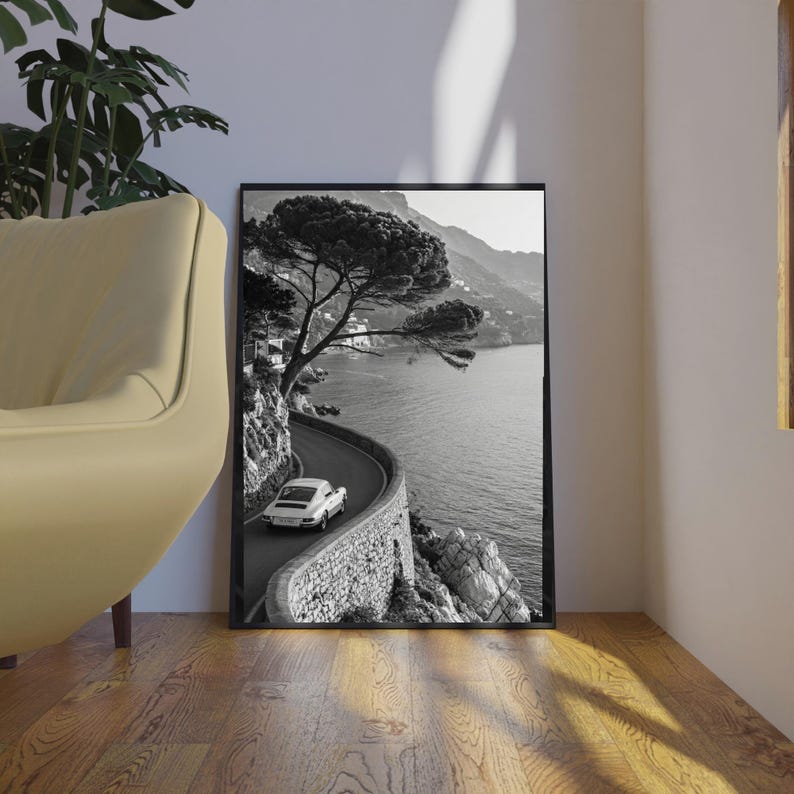 Op de afbeelding: Zwart-wit ingelijste print van een kustweg met een witte auto. De weg slingert langs de kustlijn, met aan de ene kant een stenen muur en aan de andere kant de zee. Een grote boom hangt over de weg.