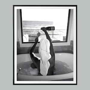 Può includere: Una persona avvolta in un asciugamano bianco è in una vasca da bagno piena di bolle. Sta versando champagne da una bottiglia in un bicchiere. La vasca da bagno è davanti a una finestra con vista sul mare.
