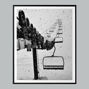 Puede incluir: Fotografía en blanco y negro de un telesilla en una tormenta de nieve. Las sillas del telesilla están suspendidas de un cable, y la nieve cubre la maquinaria del telesilla. La imagen está enmarcada en un borde negro.
