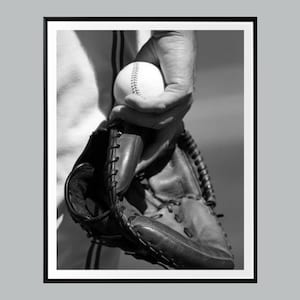 May include: A black and white photograph showing a baseball held in a leather baseball glove. The ball features visible stitching along the seams, and the glove displays detailed lacing and textured leather panels, captured in a close-up view.