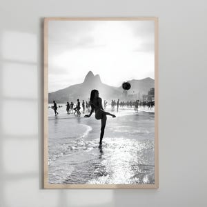 May include: Black and white framed print of a beach scene. A person is kicking a soccer ball on the shoreline, with other people in the background. Mountains and a bright sky are visible.