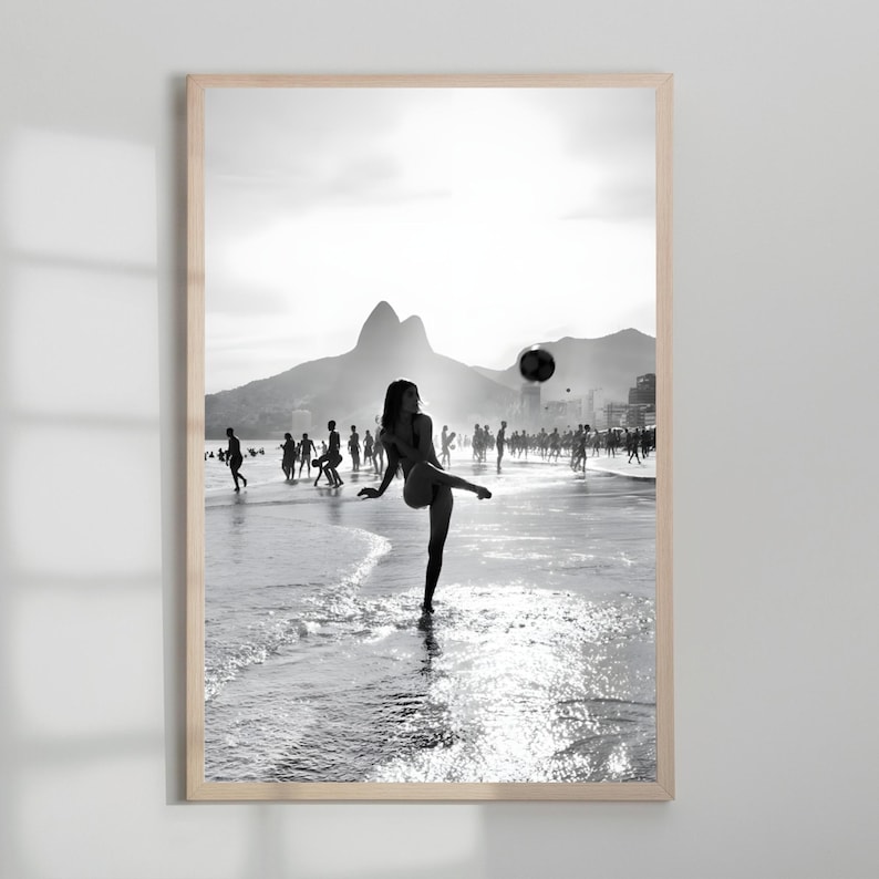 May include: Black and white framed print of a beach scene. A person is kicking a soccer ball on the shoreline, with other people in the background. Mountains and a bright sky are visible.