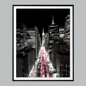 May include: Black and white cityscape print with a pink hue on the road. The image features tall buildings and skyscrapers, with a focus on the city's architecture and urban landscape. The print is framed in a black border.
