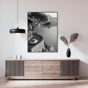 Op de afbeelding: Zwart-wit print van een kustweg met een oldtimer. De kronkelende weg volgt de kustlijn, met een stenen muur en de zee. Een boom hangt over de weg. De print wordt weergegeven boven een houten kast.