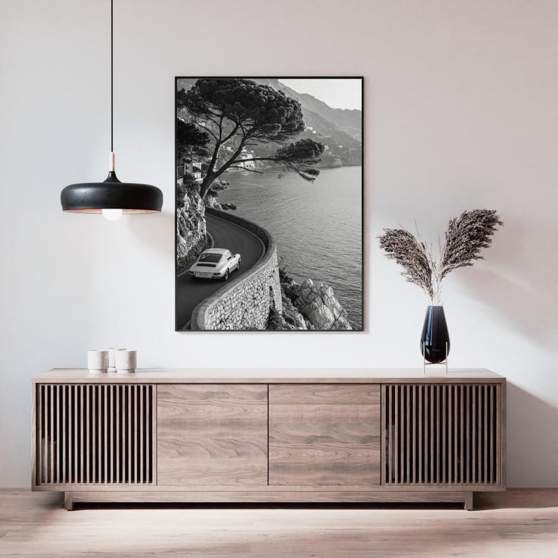 Op de afbeelding: Zwart-wit print van een kustweg met een oldtimer. De kronkelende weg volgt de kustlijn, met een stenen muur en de zee. Een boom hangt over de weg. De print wordt weergegeven boven een houten kast.