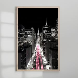 May include: A framed black and white photograph of a city street at night. The street is lined with tall buildings, and the road is illuminated with pink lights. The Empire State Building is visible in the distance.