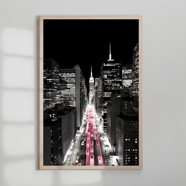 May include: A framed black and white photograph of a city street at night. The street is lined with tall buildings, and the road is illuminated with pink lights. The Empire State Building is visible in the distance.
