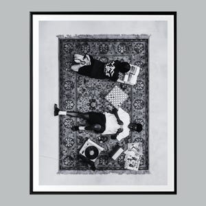 May include: Monochrome framed print showing two people on a patterned rug. A chessboard, books, and a record player are present. One person wears a black shirt with white text, the other a white shirt.