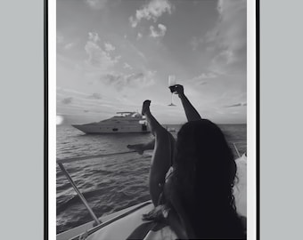 May include: Black and white photograph of a person on a boat, holding a champagne glass up in the air. A yacht is in the background on the water, with a cloudy sky above.