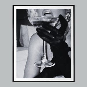 May include: Black and white photograph of a person holding a martini glass. The person is wearing a black glove and a large, ornate earring. The glass is filled with a clear liquid.