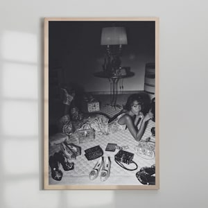 Op de afbeelding: Zwart-wit ingelijste foto van een vrouw die op een bed ligt, omringd door schoenen, handtassen en een rekenmachine. Een lamp en een bijzettafel staan op de achtergrond. De afbeelding heeft een vintage uitstraling.