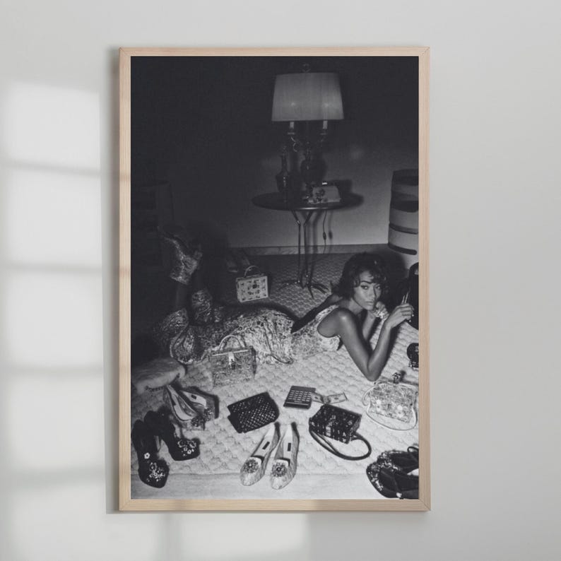 Op de afbeelding: Zwart-wit ingelijste foto van een vrouw die op een bed ligt, omringd door schoenen, handtassen en een rekenmachine. Een lamp en een bijzettafel staan op de achtergrond. De afbeelding heeft een vintage uitstraling.