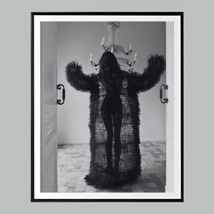 May include: Black and white photograph of a person wearing a long, ruffled black coat with arms outstretched. The person is standing in a doorway, with a chandelier visible in the background. The image is framed in a black border.
