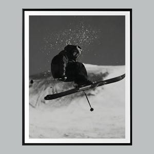 May include: Black and white photograph of a skier mid-air, captured in a dynamic action shot. The skier is wearing dark winter gear and skis, with snow particles suspended in the air. The image is framed in a black border.