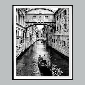 May include: Black and white framed print of a Venetian canal scene. A gondola with a striped-shirted gondolier navigates the waterway under a stone bridge. Buildings line the canal's edges, creating a classic European cityscape.