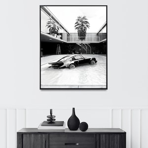K&ouml;nnte beinhalten: Schwarzwei&szlig;foto eines klassischen Sportwagens, der teilweise in einem Schwimmbad versunken ist. Das Auto befindet sich im Vordergrund, im Hintergrund ist eine moderne Architektur mit einer Palme zu sehen.