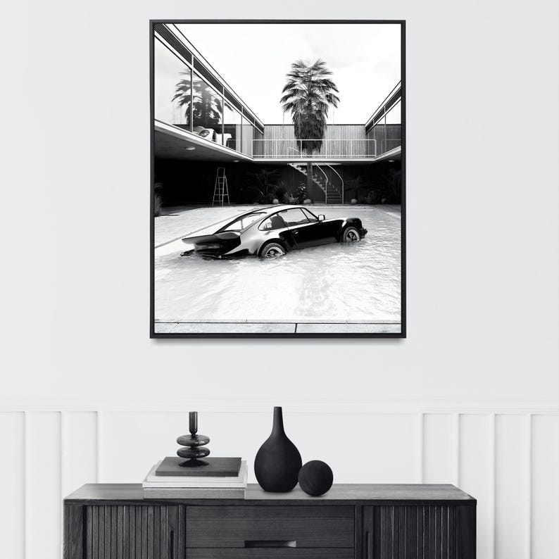 K&ouml;nnte beinhalten: Schwarzwei&szlig;foto eines klassischen Sportwagens, der teilweise in einem Schwimmbad versunken ist. Das Auto befindet sich im Vordergrund, im Hintergrund ist eine moderne Architektur mit einer Palme zu sehen.