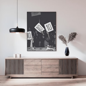 May include: Black and white framed photograph of a crowd holding up signs with the number 10. The photograph is hanging above a wooden cabinet with a black pendant light and a vase of dried plants.