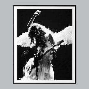 May include: A monochrome image of a person playing an electric guitar on stage, wearing a white dress and angel wings. The person has one arm raised. The guitar is white. The background is dark.