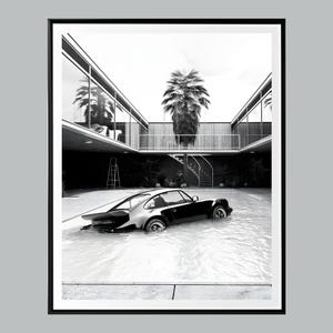 May include: A black and white photograph of a classic sports car partially submerged in a swimming pool. The car is in a modern, minimalist courtyard with a palm tree and a staircase.