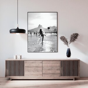 May include: Black and white framed print of a beach scene. A person is kicking a ball on the shoreline, with mountains in the background. The print is displayed above a wooden cabinet with a black pendant light.