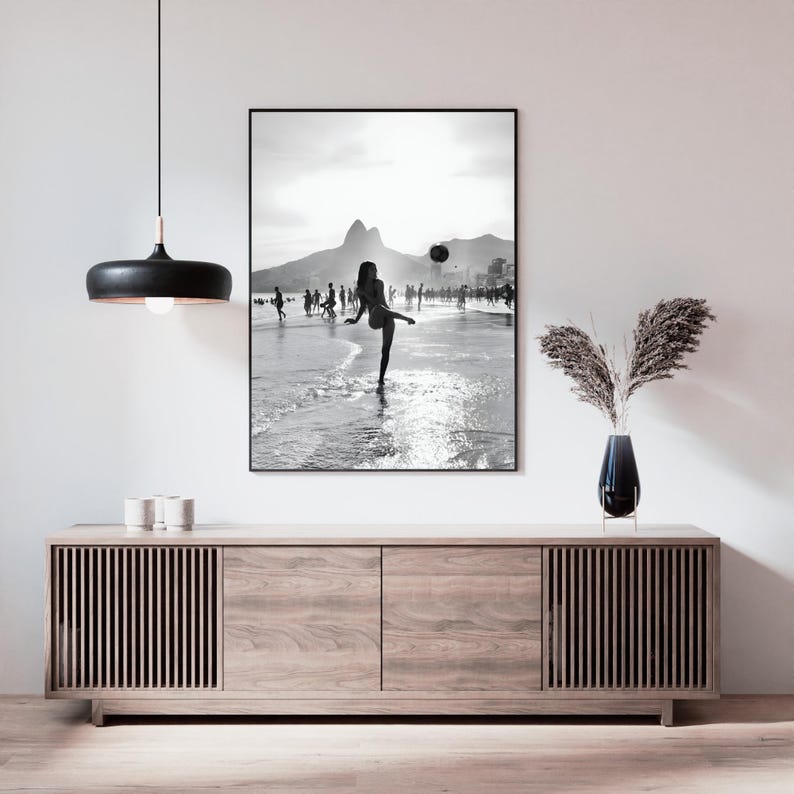 May include: Black and white framed print of a beach scene. A person is kicking a ball on the shoreline, with mountains in the background. The print is displayed above a wooden cabinet with a black pendant light.