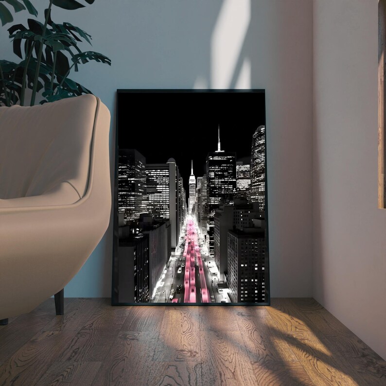 May include: Black and white framed print of a city street at night, with a pink glow down the center. The Empire State Building is visible in the distance. The print is displayed on a wooden floor.