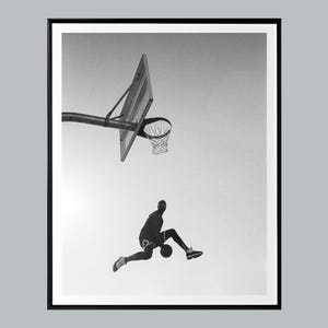 May include: Monochrome framed print showing a basketball player in mid-air, about to dunk the ball. The hoop and backboard are in the background against a bright sky. The image is a silhouette.