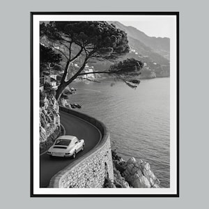 May include: Monochrome framed print featuring a classic car on a winding coastal road. The road is bordered by a stone wall and the sea. A large tree with overhanging branches frames the scene, creating a scenic view.