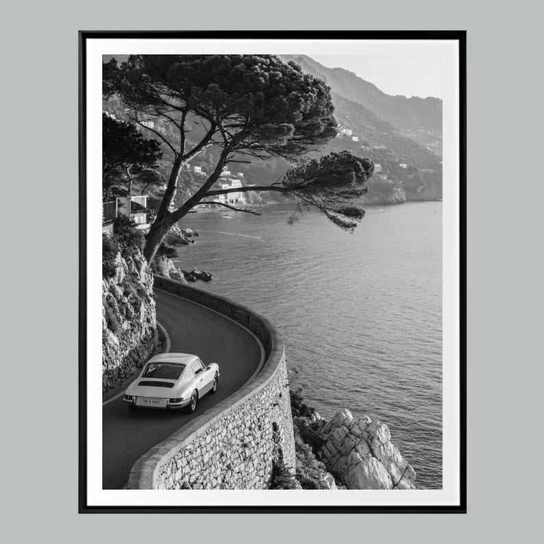 Op de afbeelding: Zwart-wit ingelijste print van een oldtimer die over een kustweg rijdt. De weg slingert langs de zee, met een stenen muur aan de ene kant en een rotsachtige klif aan de andere kant. Een grote boom hangt over de weg.