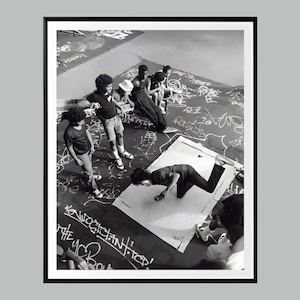 Könnte beinhalten: Schwarz-Weiß-Fotografie einer Gruppe von Personen im Freien. Einige stehen, andere sitzen, und eine Person führt eine Breakdance-Bewegung auf einem großen weißen Blatt aus. Der Boden ist mit Graffiti bedeckt.
