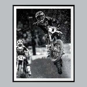 Puede incluir: Imagen en blanco y negro de un piloto de motocross en el aire, realizando un salto en una moto de cross. El piloto lleva casco y equipo de protección. Otro piloto es visible en el fondo. Gotas de agua suspendidas en el aire.