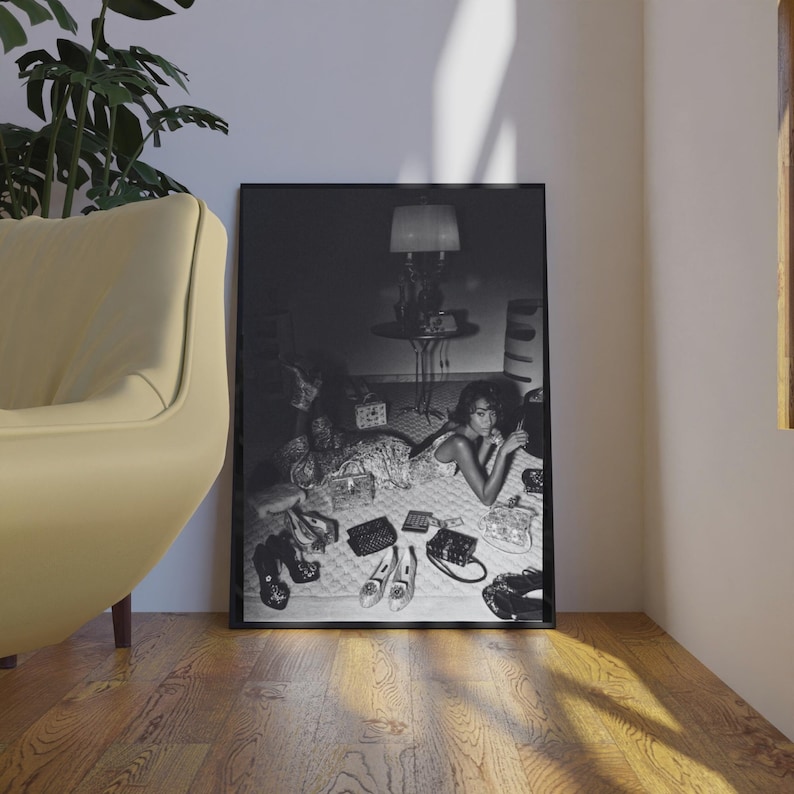 Op de afbeelding: Zwart-wit ingelijste print met een vrouw omringd door handtassen, schoenen en accessoires. De afbeelding is een foto in vintage stijl met een lamp en een bijzettafel op de achtergrond. De sc&egrave;ne speelt zich af op een oppervlak met patronen.