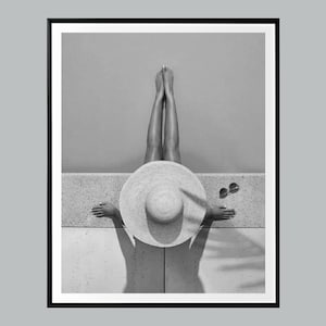 May include: Black and white photograph of a person relaxing outdoors. The person is wearing a large sun hat and sunglasses, with their legs raised. The image is framed in a black border.
