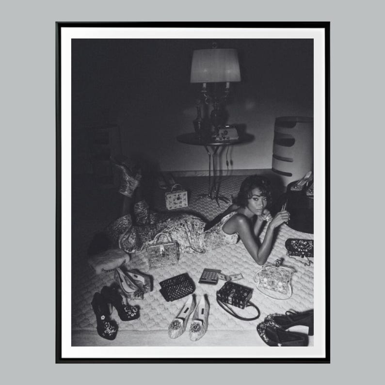 Op de afbeelding: Zwart-witfoto van een vrouw die op een bed ligt, omringd door diverse handtassen, schoenen en accessoires. Een lamp en een bijzettafel zijn op de achtergrond te zien. De afbeelding heeft een mode- of lifestyle-thema.
