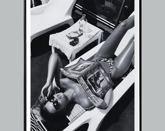 May include: Black and white photograph of a person reclining on a white lounge chair, reading a magazine. The person is wearing sunglasses and a leopard print bikini bottom. A small table with a bottle and other items is nearby.