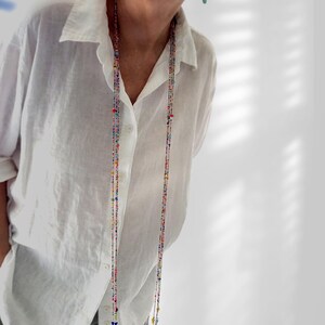 May include: A long, multi-strand necklace featuring tiny, vibrant, multi-coloured glass beads in shades of red, blue, yellow, and green. The delicate, thin strands hang vertically, creating a layered effect against a plain white button-down shirt.