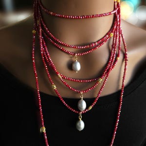 May include: A multi-strand necklace featuring numerous small, faceted red beads arranged in several layers. The design includes small gold-toned metal spacers and three dangling, smooth, oval-shaped white pearls suspended from the lower strands.